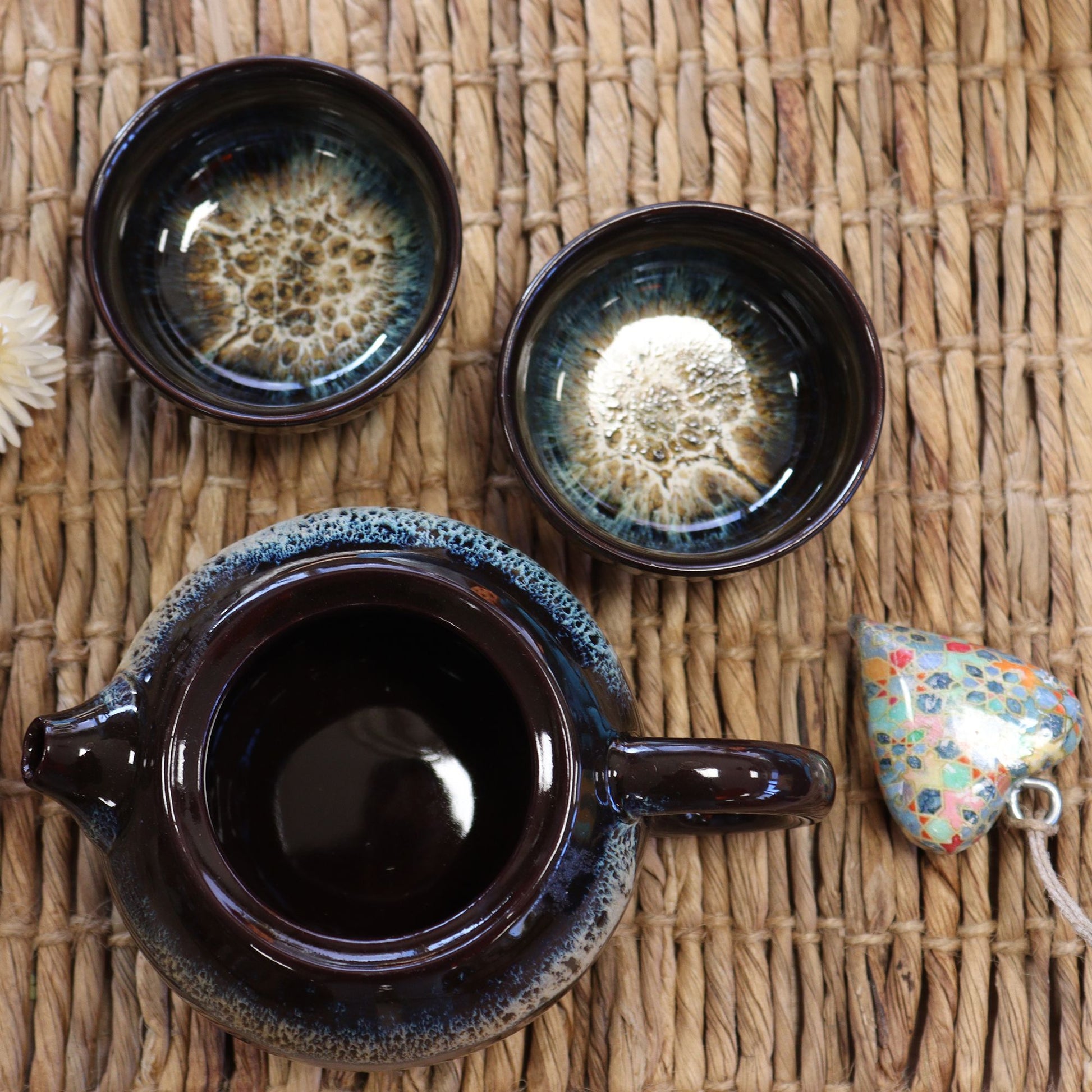 Ceramic teapot and two cups on a woven surface with a decorative item.