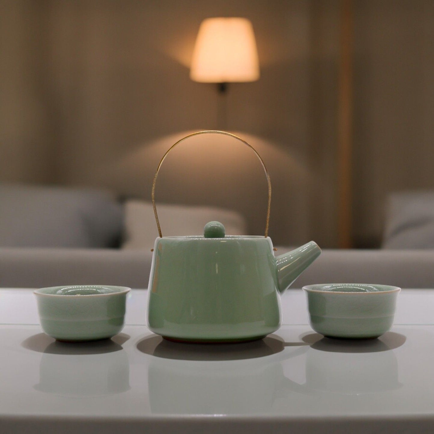Green ceramic teapot with two matching cups on a reflective surface, blurred background