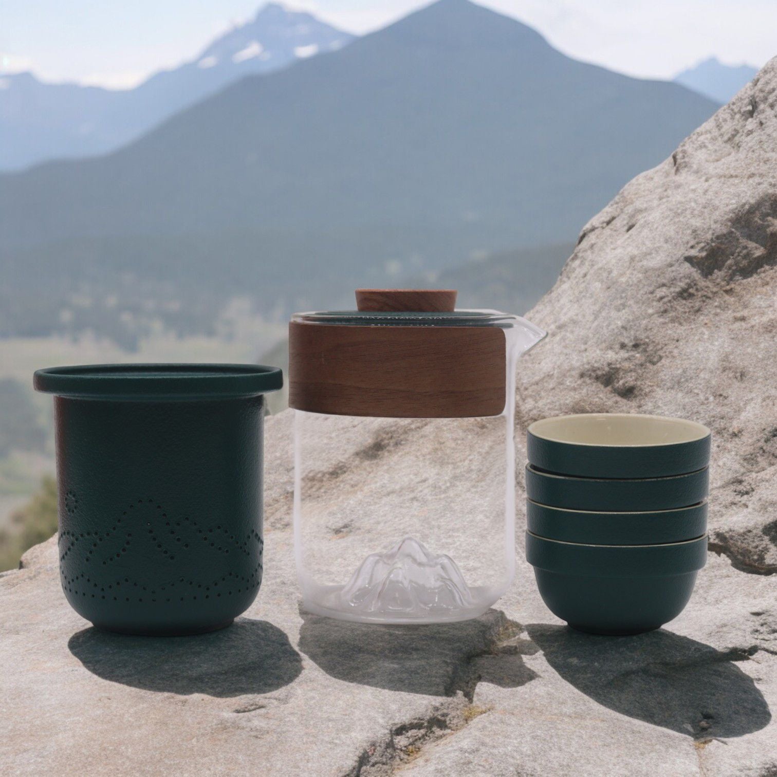Set of ceramic cups and a glass jar with wooden lid on a rocky surface with mountains in the background